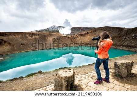 Woman photographer camera on Viti Crater of Krafla caldera geothermal formation with small lake of blue turquoise green water and mountains snow during summer
