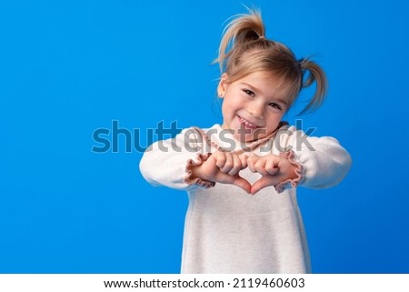 Happy little girl making heart shape by hands on blue background.
