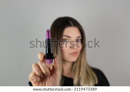 Close up photo of beautiful girl hold purple pomade. She will check new lips stick on isolated over gray background. Matte effect. Cosmetic, beauty and make up. Focus is on purple lipstick.	