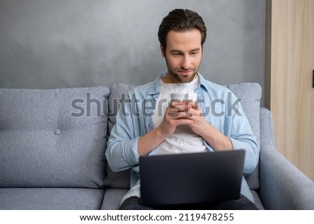 Handsome young hipster freelancer man sitting at home and looking at laptop screen, taking a break from working on laptop, holding mug and drinking coffee, enjoying working from home