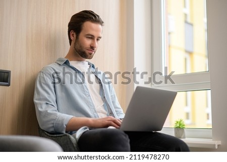 Focused determined young bearded businessman or freelancer in casual wear typing on laptop, working on project remotely, researching, writing email to client, web surfing staying at home, side view