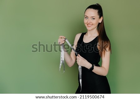 Happy slim woman in sport clothes standing with measuring tape on green background