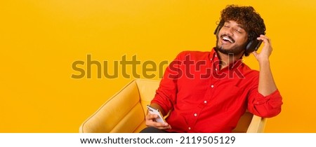 Joyful middle eastern man wearing wireless headphones holding smartphone in hand, enjoying music. Ecstatic cheerful multiracial guy listening favorite tracks, isolated on orange