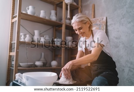 Smiling senior caucasian female master molding clay pot on pottery wheel in art studio. Small business and entrepreneurship. Home hobby, entertainment and leisure. Woman wearing work apron. Sunny day Royalty-Free Stock Photo #2119522691