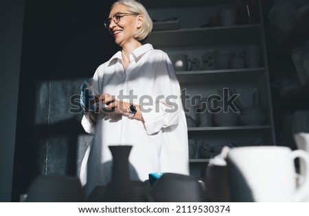Smiling mature caucasian female entrepreneur wearing work glove and looking away. Small business and entrepreneurship. Modern successful woman. Dark home art studio with pottery on shelves