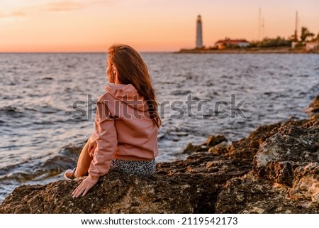 Amazing colorful sunset landscape on the sea and a young beautiful woman sitting on the rocks and admiring the lighthouse in the Crimea