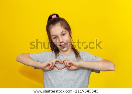 Adorable girl in gray t-shirt making a heart gesture with her fingers with a happy sincere smile isolated over yellow background. Expression of love and happiness. Mothers Day. Valentine's Day