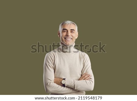 Studio portrait of senior man with happy face expression. Cheerful, smiling man wearing turtleneck sweater and wristwatch looking at camera standing with arms folded isolated on solid green background