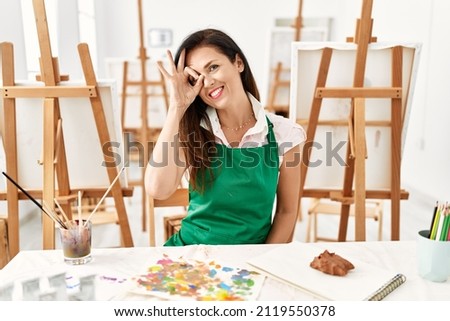 Beautiful caucasian woman at art studio smiling happy doing ok sign with hand on eye looking through fingers 