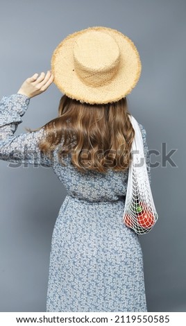 Attractive young girl in staw hat holding a mesh bag with vegetables. Picture taken from the back.Studio shot on grey background.

