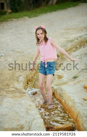 Young girl playing by natural swimming pool in Bagno Vignoni, with thermal spring water and calcium carbonate deposits, which form white concretions and waterfall. Tuscany, Italy. Royalty-Free Stock Photo #2119581926