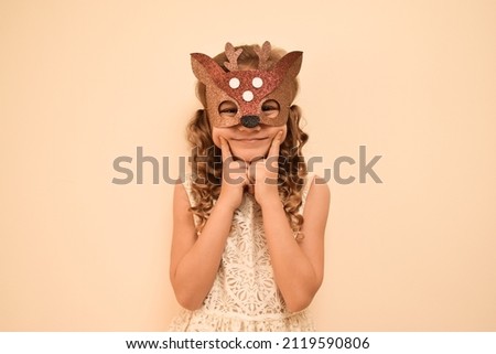 Happy child in deer costume. Little cute girl in a carnival masquerade deer mask made of shiny glitter foamiran on a beige background. The child is preparing for the masquerade. Royalty-Free Stock Photo #2119590806