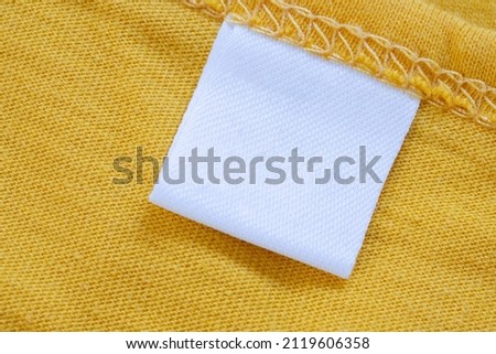White blank clothing tag label on new yellow shirt background Royalty-Free Stock Photo #2119606358