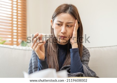 Sick, influenza asian young woman, girl headache have a fever, flu and check thermometer measure body temperature, feel illness sitting on sofa bed at home. Health care person on virus, covid-19. Royalty-Free Stock Photo #2119608002