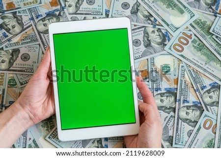 digital money, hand holds tablet with blank green touchscreen on money background or surface with american dollar banknotes. Money concept with united states of america currency and tablet computer