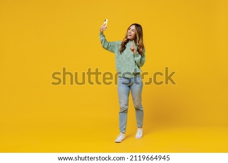 Full body young smiling woman 30s wearing green knitted sweater doing selfie shot on mobile cell phone show v-sign isolated on plain yellow color background studio portrait. People lifestyle concept