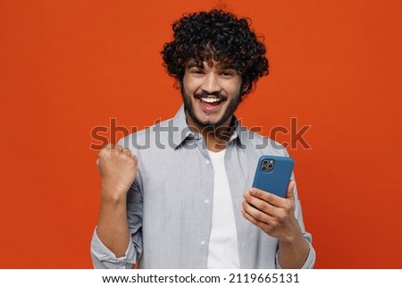 Young bearded Indian man 20s years old wears blue shirt hold in hand use mobile cell phone doing winner gesture celebrate clenching fists say yes isolated on plain orange background studio portrait Royalty-Free Stock Photo #2119665131