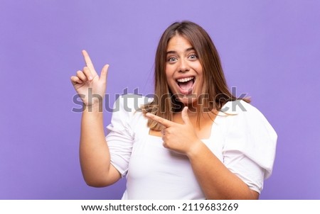 young blonde woman feeling joyful and surprised, smiling with a shocked expression and pointing to the side