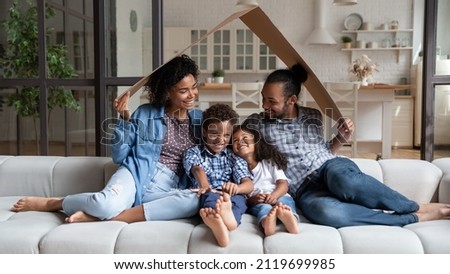 Happy affectionate young African American couple parents sitting under carton roof on comfortable couch with small adorable children son daughter, celebrating moving into own home, real estate concept Royalty-Free Stock Photo #2119699985