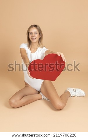 Lovely woman in white bodysuit and sneakers smiling having red paper heart in hands on background of flesh color. Celebrating and congratulating loved one with Valentine's day. Expressing love.