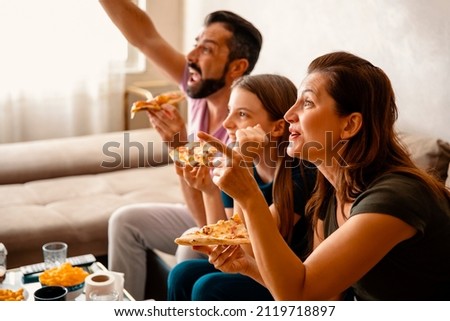 Excited Family cheering for their sport team on TV, Eating pizza in the living room at home. Fans with hands up and pizza slices in hands having fun watching favorite game on Television. Copy space Royalty-Free Stock Photo #2119718897