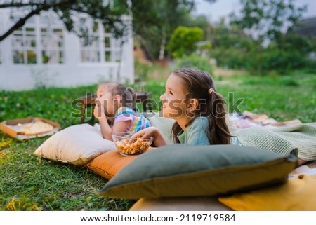 Side view of girls lying on blanket, eating homemade popcorn and watching film on DIY screen from projector. Summer outdoor weekend activities with kids. Open air cinema. Outside movie night Royalty-Free Stock Photo #2119719584