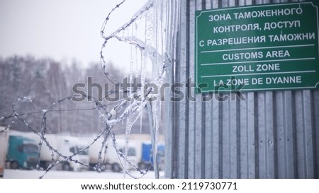 Blurred trucks behind barbed wire fence and customs area sign in Russia, same text in different languges
