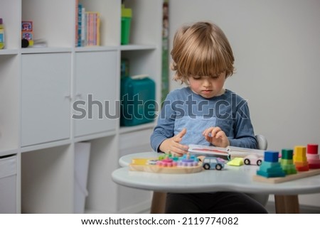 Beautiful toddler reading a book and playing with a wooden toys at home. Toddler play with a color educational toy and wooden car. Child play at the table in the baby room. Funny baby. Lifestyle. Royalty-Free Stock Photo #2119774082
