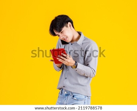 Studio shot of millennial Asian thoughtful doubtful curious male fashion model in stylish fashionable casual outfit standing call smartphone and holding red tablet on yellow background.
