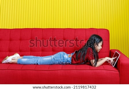 Closeup shot of Asian sleepy young primary school girl in denim jeans overalls outfit laying down holding hand cover mouth yawning while browsing surfing internet with touchscreen tablet computer.