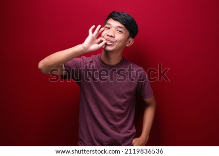 Portrait of asian man making delicious hand gesture, close up head and shoulder portrait against red background