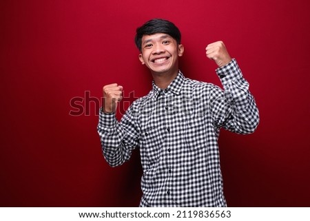 asian man wearing plaid shirt with happy expression showing victory, on red background