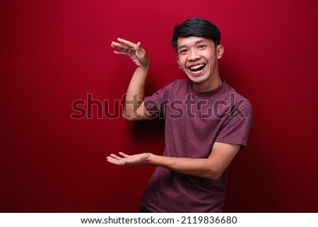 asian man wearing tshirt over red background gesturing with hands showing big and large size sign, measure symbol. smiling looking at the camera. measuring concept.