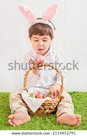 a small child in rabbit ears sits on the floor with a basket of colored eggs