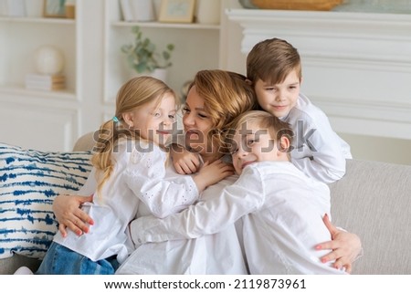 We love you. Smiling young woman, kids hug mom while sitting on sofa in living room at home. Happy caucasian family enjoying a day together in a bright room