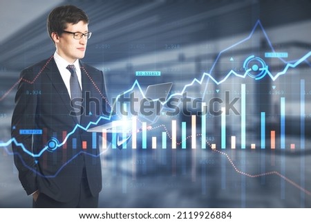 Attractive young european busiessman holding laptop with creative glowing forex chart on blurry office interior background. Trade, financial investment and technology concept. Double exposure
