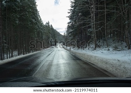 A shot on the move from behind the windshield of an electric car with snow-covered mountains. Cold cloudy winter day. POV first person view shot on a asphalted mountain road. Selective focus.