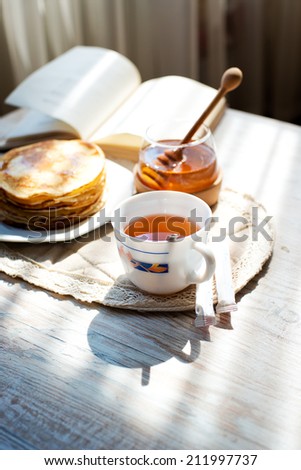 A cup of hot tea with honey pancakes and a book by the window