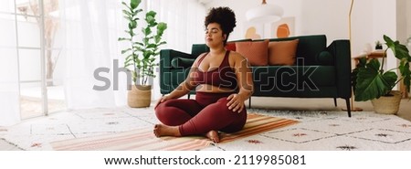 Plus size woman sitting cross legged and closed eyes in living room. Pretty female in fitness wear practicing meditation at home. Royalty-Free Stock Photo #2119985081