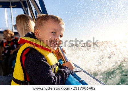 Portrait of cite little blond happy excited smiling caucasian boy wear lifevest enjoy sailing on motor boat sea against blue sky and water splash wave sun backlit. Summer travel vacation recreation Royalty-Free Stock Photo #2119986482