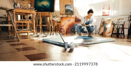 Artist making a blue painting on a canvas. Female painter touching her painting while squatting on the floor in her art studio. Creative young woman working on a new project. Royalty-Free Stock Photo #2119997378