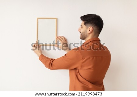 Modern Home Interior And Domestic Decor. Portrait of smiling man hanging wooden photo picture frame on the white wall. Casual guy holding showing empty poster for design mockup, blank free copy space