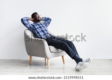 Peaceful young black man relaxing in armchair against white studio wall, free space. Full length of African American guy enjoying lazy weekend morning, resting with hands behind head Royalty-Free Stock Photo #2119998551