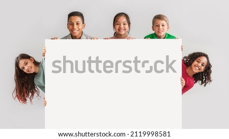 Happy Diverse Kids Hiding Behind Empty White Board Smiling To Camera Posing Over Gray Studio Background. Mixed Boys And Girls Advertising Your Text On Blank Poster. Panorama, Mockup Royalty-Free Stock Photo #2119998581