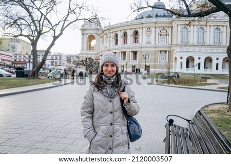 Smiling woman with a backpack standing on the background of the house in a classic style. Woman walking in the city center near the theater.