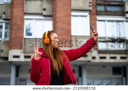 a girl in her early twenties in a red jacket with headphones takes a self photo in front of buildings