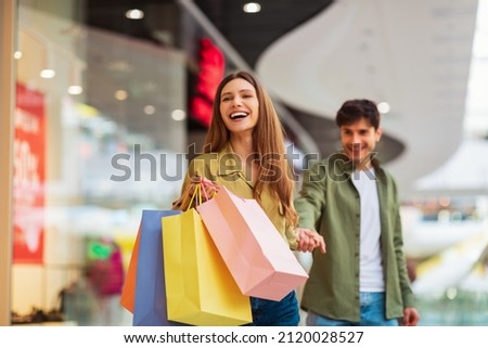 Shopping Together. Joyful Wife Carrying Shopper Bags Pulling Husband Holding His Hand And Laughing Enjoying Sales Weekend In Modern Mall. Shopaholism And Commerce. Selective Focus Royalty-Free Stock Photo #2120028527