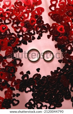 Gold wedding rings and red heart confetti on pastel pink background. Flat lay.
