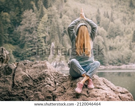 A woman sits on a large rock with her hands folded over her head and her face hidden under her hair near a lake in the forest. Joking photo Royalty-Free Stock Photo #2120046464