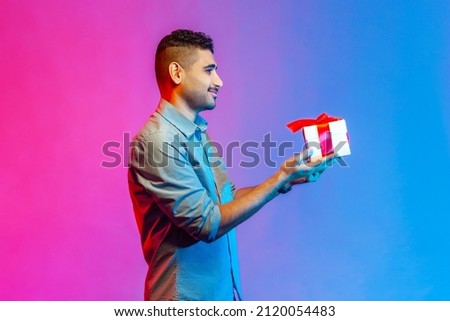 Side view of positive delighted man in shirt standing giving gift box, smiling looking friendly and generous, charity. Indoor studio shot isolated on colorful neon light background.